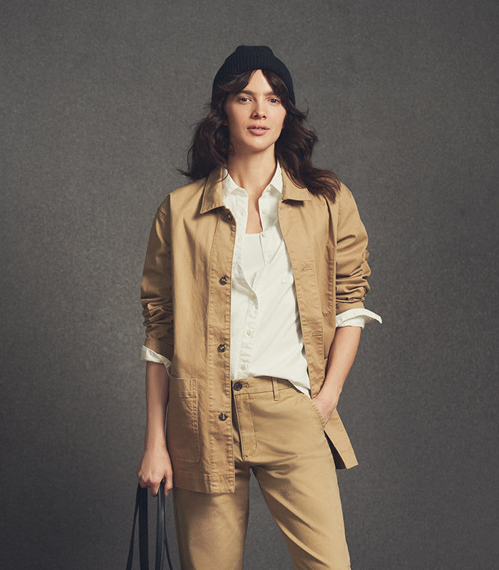Woman wearing a beige jacket and pants with a white shirt, holding a black bag against a gray background