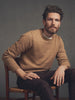 Man wearing a brown sweater sitting on a chair against a gray background