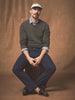 Man sitting on a stool wearing a green sweater, blue pants, and a cap against a brown background